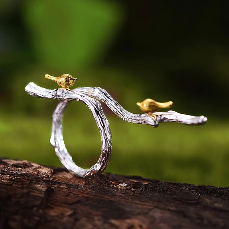 Silver ring with gold birds on a branch against a blurred green background
